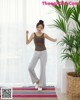 A woman standing in front of a window wearing a brown tank top and gray pants.