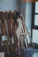 A naked woman standing in front of a wooden easel.